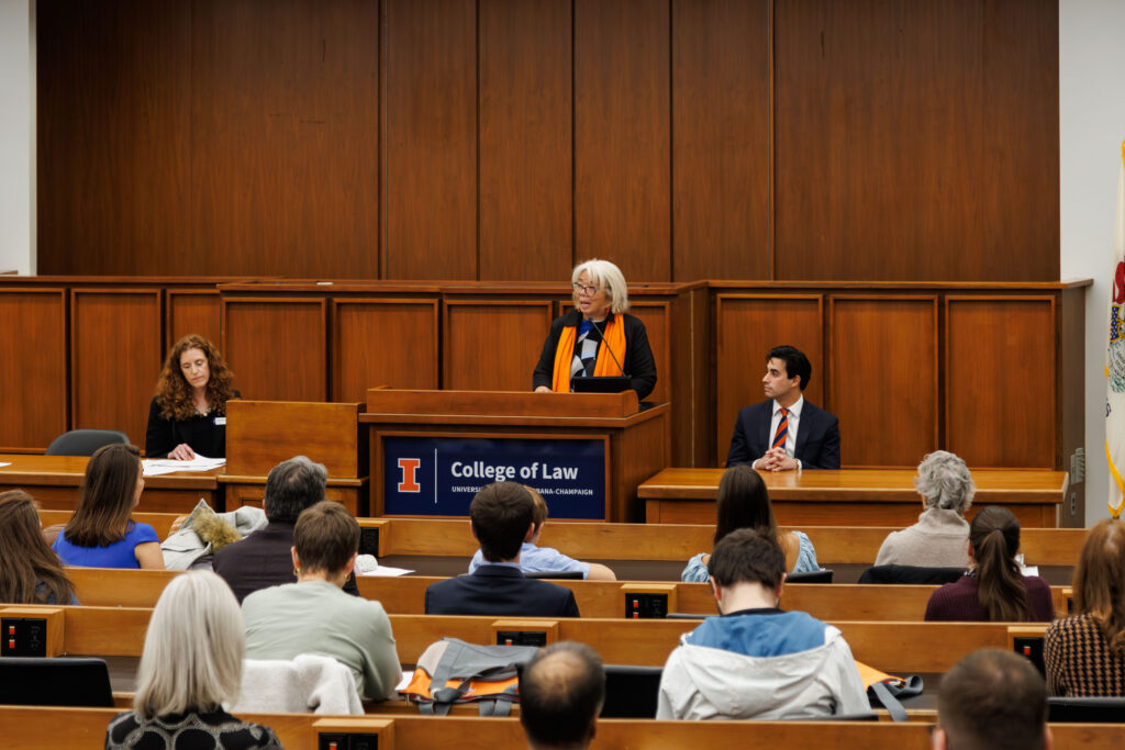 Dr. Amy Santos speaks at a podium, with Colleen Murphy to her left and Jake Sherkow to her right.